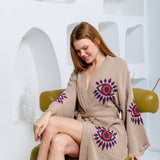 A woman wearing a beige cotton robe with purple and red eye motifs sits on a green armchair against a white textured wall with arched recesses.