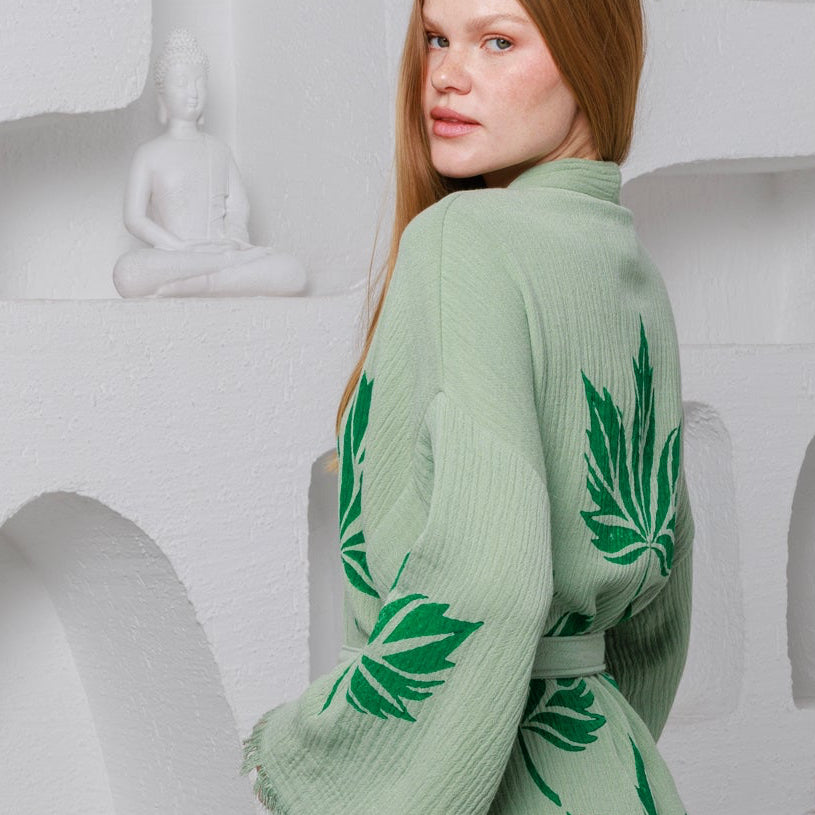 A woman stands in a white room with sculptural wall niches, wearing a light sage green kimono-style robe with large green leaf prints. A mustard yellow chair, a black lantern, and a potted trailing plant are nearby.